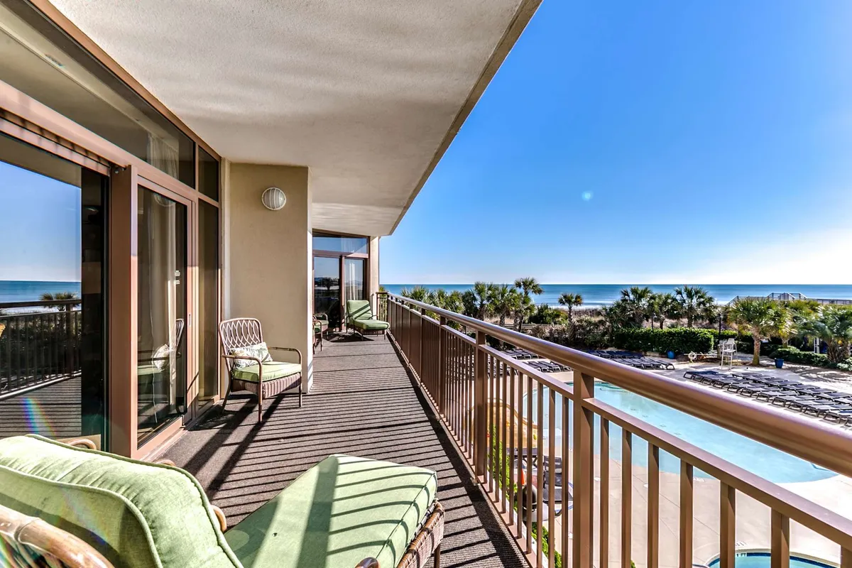 An oceanfront balcony furnished with comfortable seating, overlooking the pool area, palm trees, and the vast ocean beyond. The setting is ideal for morning coffees, sunset toasts, and relaxed conversations set against a mesmerizing seascape.