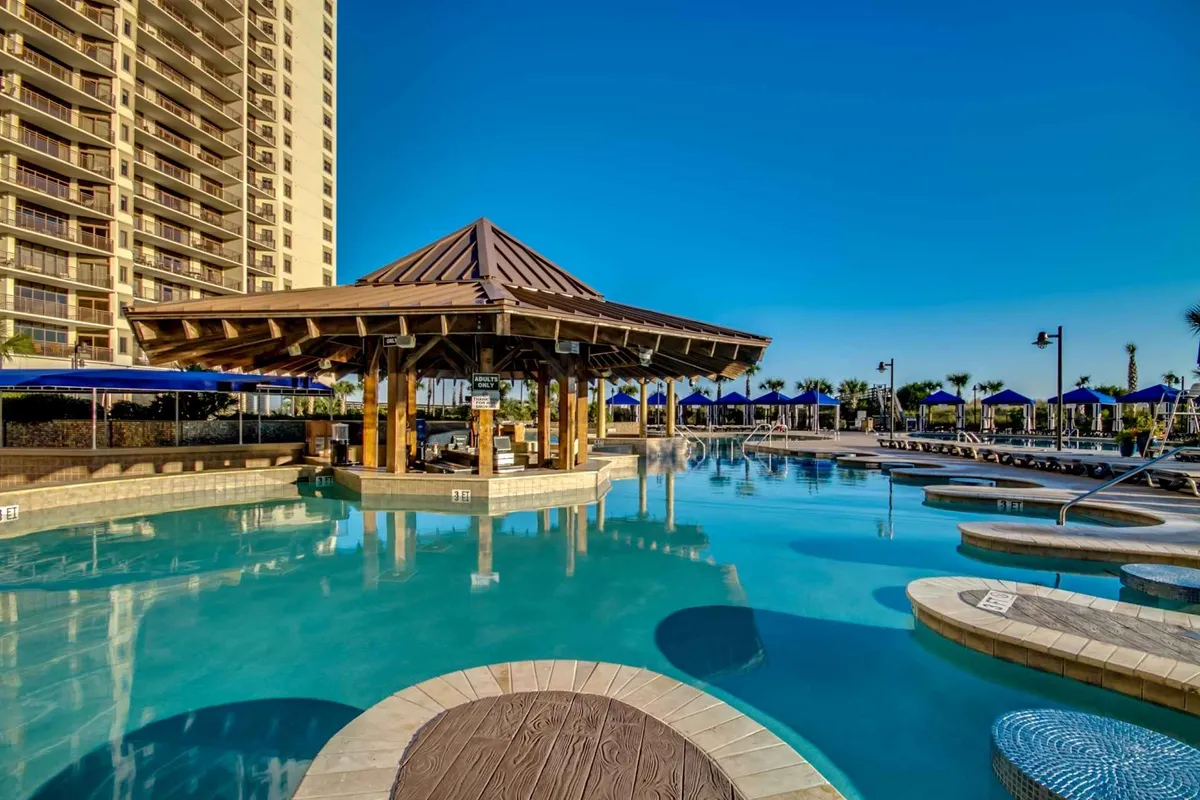 An inviting pool area with a wooden pavilion, blue umbrellas, and lounge chairs, creating a cheerful and leisurely environment. The setting hints at sunshine-filled days spent swimming, socializing, and relaxing poolside—a central feature for families staying at the resort.