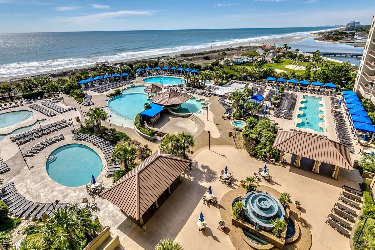 A vibrant exterior view of the resort's main swimming pools surrounded by palm trees and lounge chairs, with a beachfront in the background. The scene is lively and inviting, capturing the essence of resort leisure and social activity.