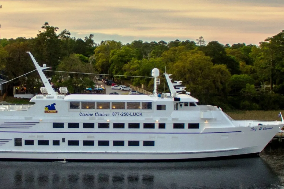 The photo shows the exterior of a casino cruise boat on the water, surrounded by trees and with scenic views. Groups of people can be seen socializing on deck, evoking the sense of adventure and festivity that comes with a special outing.