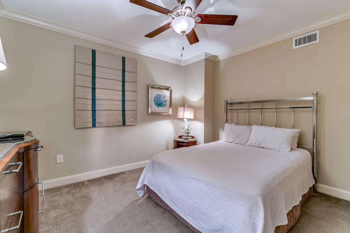 A tranquil bedroom featuring a plush bed dressed in soft linens, a ceiling fan overhead, a bedside lamp for evening reading, and tasteful artwork on the wall. The space appears inviting and designed for relaxation, with subtle decor touches that enhance comfort.