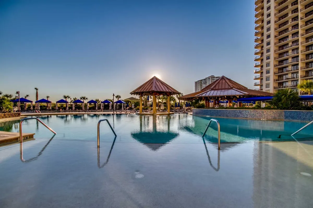 A tranquil scene featuring a sparkling swimming pool with thatched-roof cabanas, sunset reflections on the water, and surrounding palm trees, with the high-rise resort building in the background. The setting feels exclusive and serene.