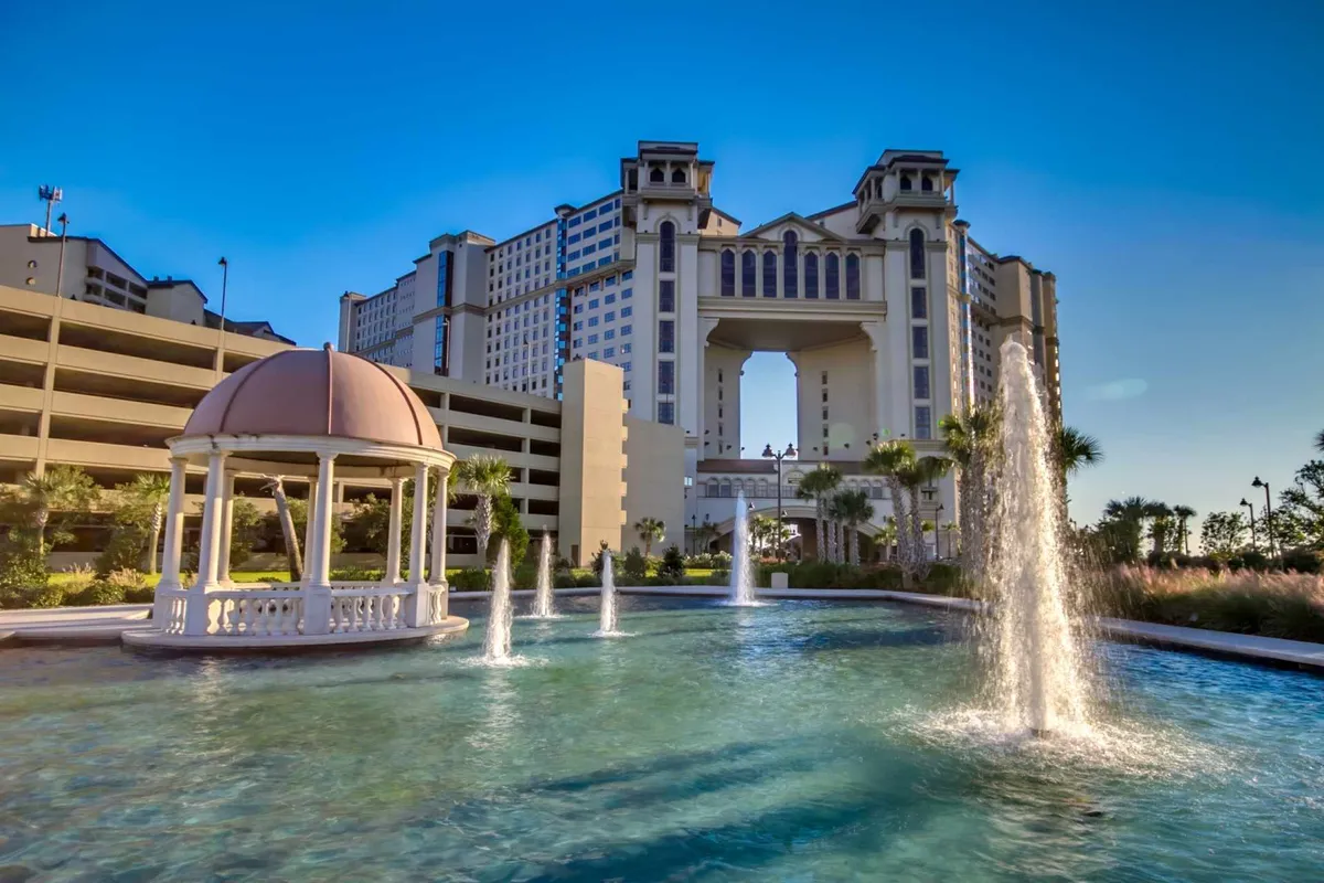 A grand, ornate gazebo sits beside serene fountains and an expansive pool area, framed by a luxury resort building. The sunset sky is reflected in the water, creating a tranquil yet vibrant setting ideal for socializing, relaxing, or celebrating with friends.