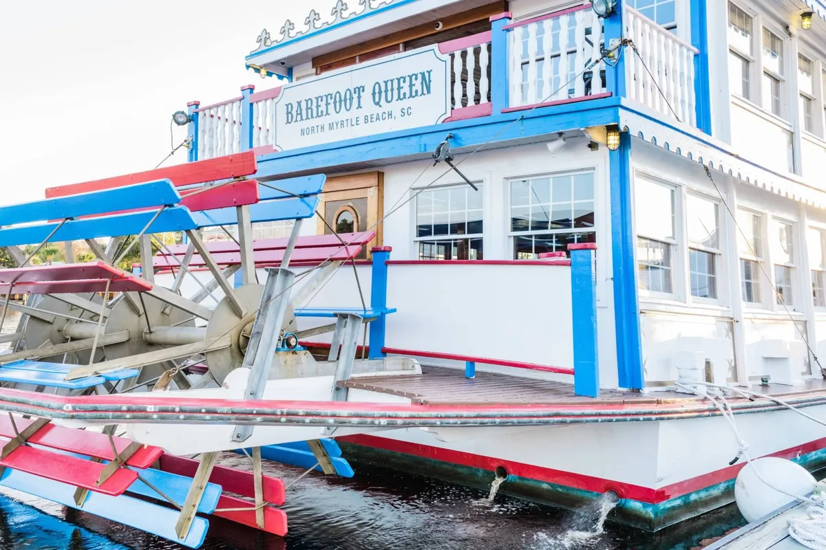 A brightly painted paddlewheel boat named 'Barefoot Queen' is docked by the water. The boat is bustling with energy, promising live music, dining, and scenic sunset tours. The vivid exterior and festive signage create a lively atmosphere ideal for evening adventures.
