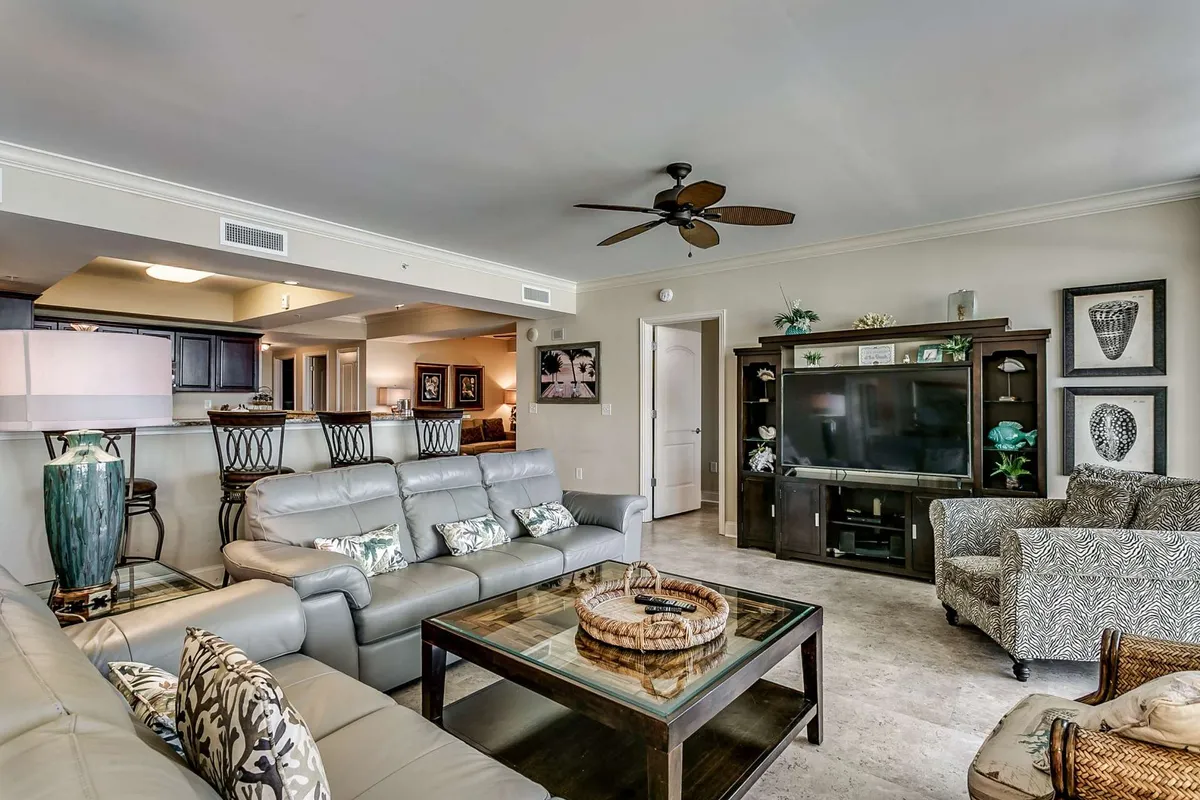 A warm, inviting den/media room with plush leather sofas, decorative pillows, a sleek coffee table, and an entertainment unit topped with a TV. The ceiling fan adds comfort, and the overall decor encourages relaxation and socializing.