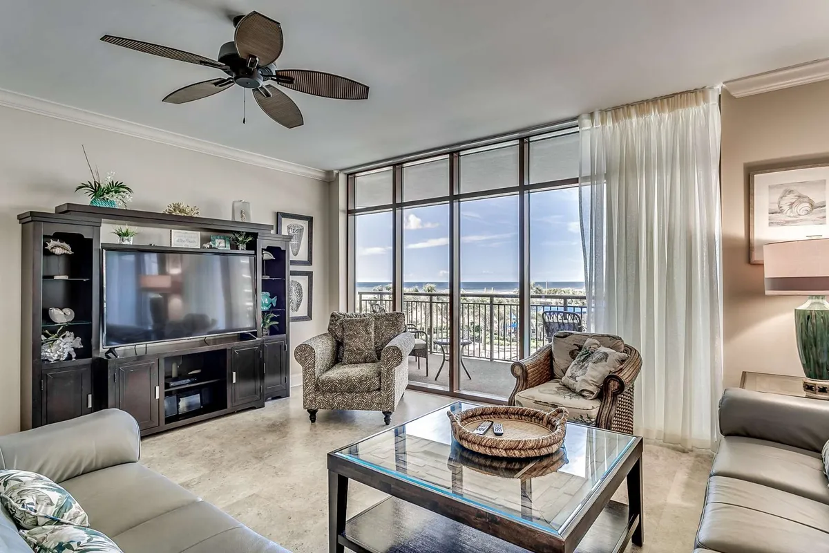 A spacious, cozy living area featuring comfortable seating arranged around a TV unit, with a large window offering a striking ocean view. The room is styled for relaxation and socializing, inviting groups to gather and enjoy entertainment or conversation while basking in the natural light and scenery.