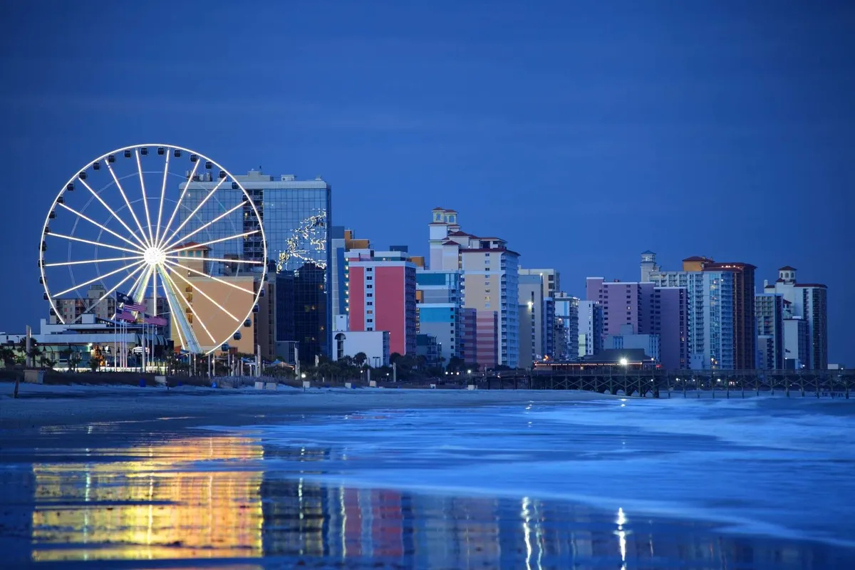 A vibrant exterior scene featuring a colorful waterfront with a ferris wheel, lively amusement buildings, and their reflections shimmering on the water. The atmosphere is bustling and energetic, inviting families to enjoy amusement rides, evening strolls, and Myrtle Beach nightlife.