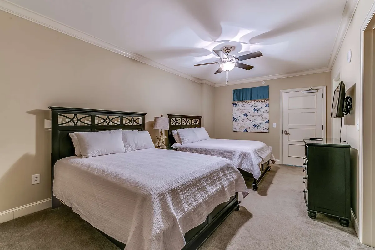 A cozy bedroom with two beds, decorative curtains framing the windows, a ceiling fan for comfort, and a TV positioned for easy viewing. Ample natural light fills the room, creating a bright and welcoming space ideal for relaxation or catching up on favorite shows.