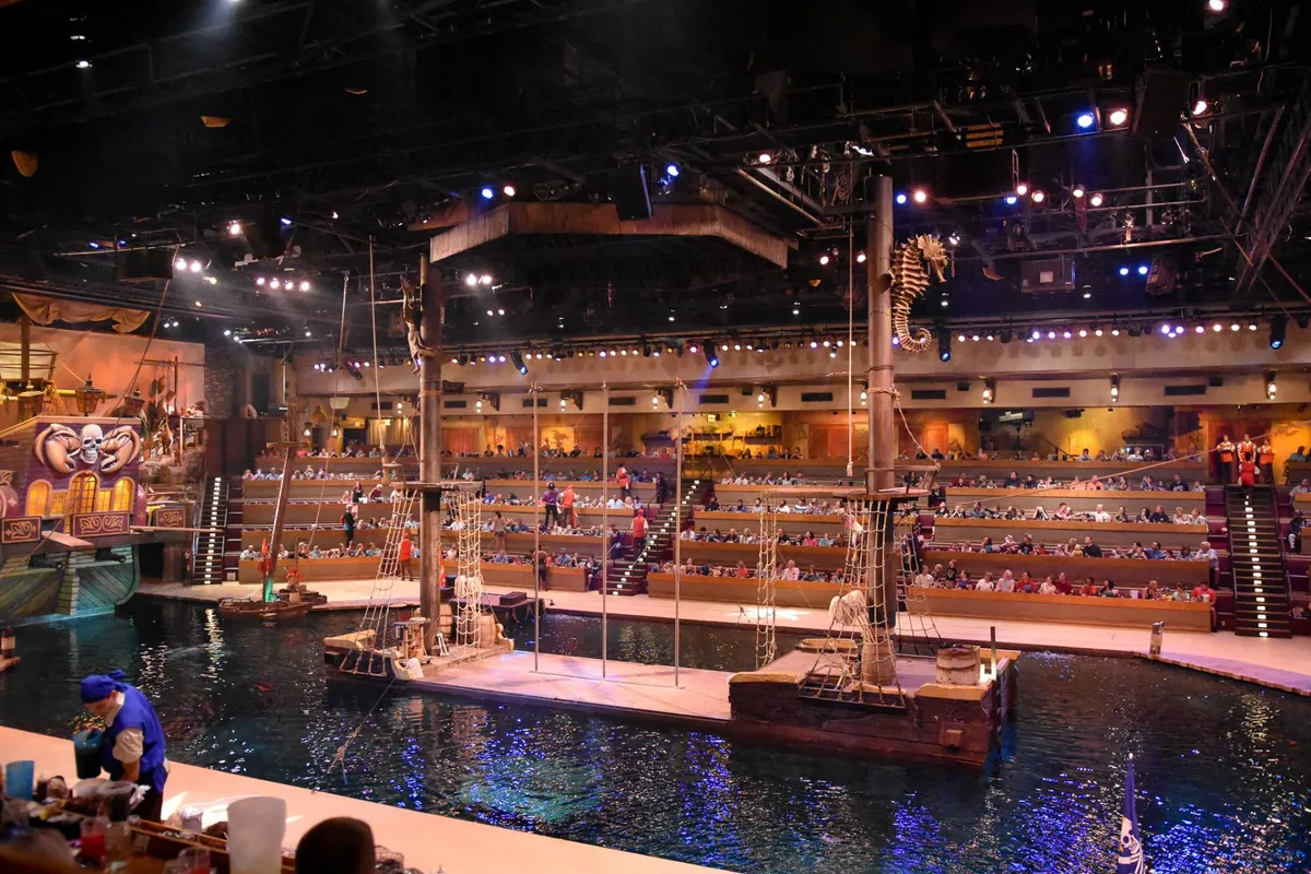 This image captures an exciting live performance at the Pirate’s Voyage Dinner & Show, featuring a large ship structure at the center, a lively audience in the stands, and dramatic lighting reflecting off the water. The image conveys the immersive and festive atmosphere of the show, with vibrant costumes and interactive moments.